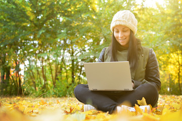 Girl in hipster with laptop in autumn park. A woman in a cap using a laptop while sitting on fallen leaves. Freelancer in the hat uses remote communication technology. Remote work. Free space