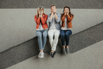 Friends sitting on parapet and closing faces with hands. Copy space on right side