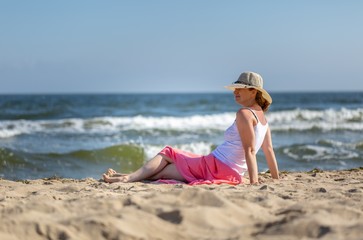 Beautiful blonde woman in hat and skirt sitting on sea shore.