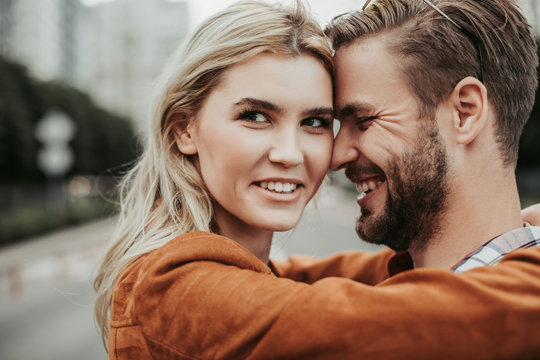 Happy twosome embracing at street. She is looking at camera with bright smile while he touching her with forehead