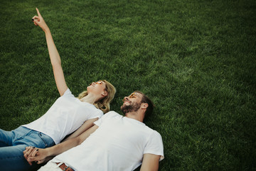 Top view of cute happy twosome lying on lawn and holding hands. Woman pointing up. Copy space on right side