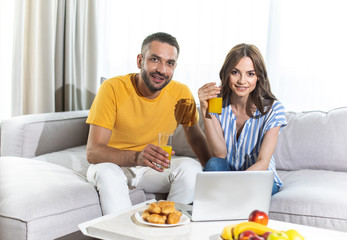 Beautiful woman and smiling man is drinking orange juice and looking at laptop while sitting on sofa