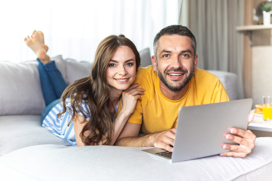 Cheerful man and woman are lying on couch and holding laptop while smiling