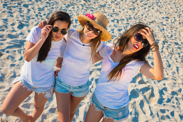 Tres chicas sexys de blanco en la playa sobre la arena riendo y haciendo selfies.
