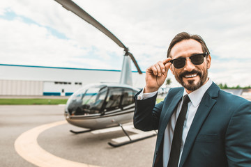 Positive smile. Attractive young businessman smiling happily while touching his glasses and standing on the helicopter platform