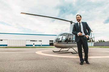 Calm handsome elegant man in dark suit feeling confident and looking into the distance while standing on the helicopter platform - Powered by Adobe
