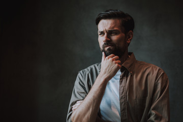 Portrait of pensive unshaven male keeping chin with hand and grimacing face. He isolated on dark background