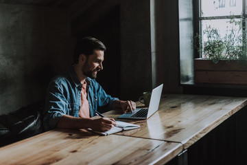Side view positive unshaven male making notes while using contemporary laptop during work at table