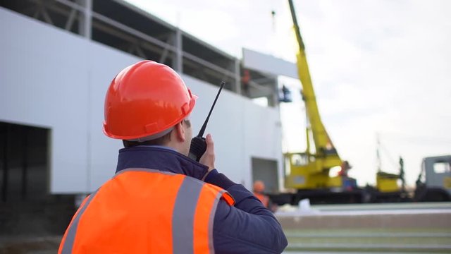 Male engineer, builder, foreman, worker in orange a uniform and hard hat using a walkie talkie giving instructions at a construction site. installation of sandwich panels of unfinished building