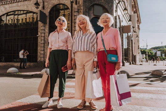 Full-length photo of three laughing older women holding shopping bags while standing on pedestrian crossing