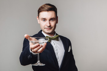 Elegant young man in black suit and bow tie holding a drink and making toast gesture isolated on grey background. New year's eve. Offering a drink