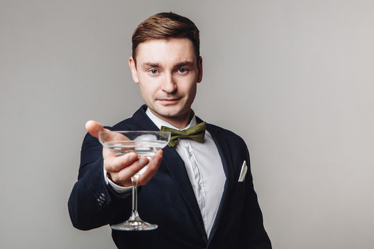 Elegant Young Man In Black Suit And Bow Tie Holding A Drink And Making Toast Gesture Isolated On Grey Background. New Year's Eve. Offering A Drink