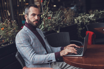 Serious person. Calm thoughtful bearded man sitting at the table with modern laptop on it and...