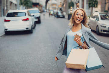 Waist up portrait of beautiful middle-aged lady in glasses holding cup of coffee and shopping bags while looking at camera and laughing