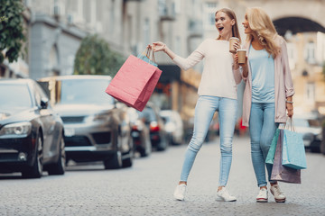 Full length portrait of beautiful girl and her mother holding shopping bags and cups of hot drink. Women looking away and smiling as if they saw something exciting