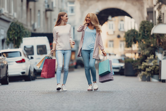 Full length portrait of charming young lady chatting with mother outdoors. They carrying colorful shopping bags while looking at each other and smiling