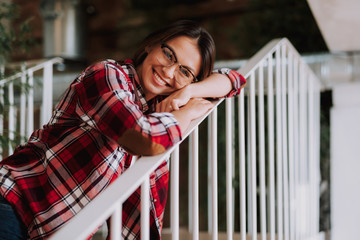 Portrait of charming woman in plaid shirt putting head on hand while looking at camera and smiling