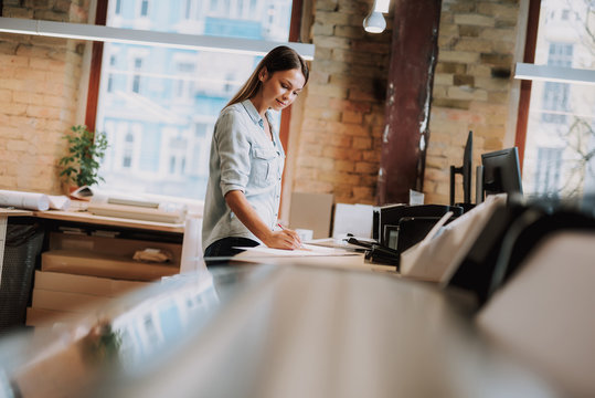 Side view portrait of charming girl in blue shirt making notes while standing near table with printer