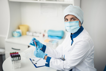 Fototapeta premium Professional laboratory research in healthcare system. Female facility assistant looking at camera while holding test tube of blood in lab office