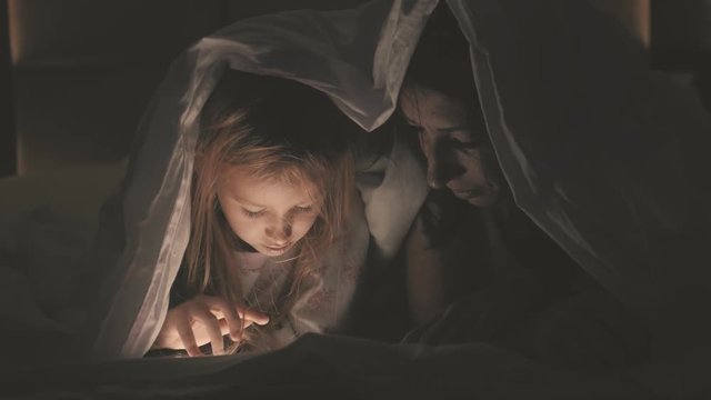 Young woman mom and her daughter watching interesting film together on tablet and laugh under the blanket. Funny mom and lovely child are having fun with tablet. Happy loving family.