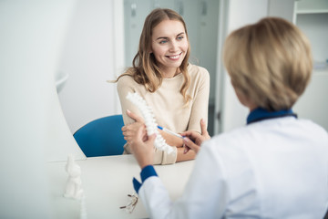 Concept of professional consultation in healthcare system. Waist up portrait of female doctor showing medical spine model to smiling young lady in clinic