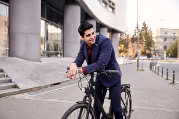 one young man, 20-29 years old, wearing suit, looking sideways,  smiling. posing on fancy city bicycle, leaning to front handlebar. upper body shot. modern architecture building behind.
