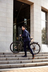 one young handsome man, wearing suit, pulling bicycle up on stairs steps, modern architecture building.
