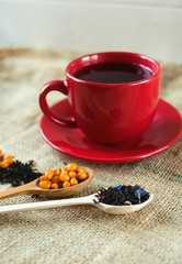 Vitaminic healthy sea buckthorn tea in red cup with fresh raw sea buckthorn berries and black tea in wooden spoons over sackcloth background. warm winter  organic drink, health food concept 