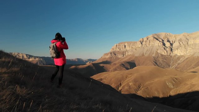 Aerial view of a girl traveler with a backpack and a camera takes pictures in the mountains at sunset. Girl photographer from the back. The camera passes by and flies to the mountains
