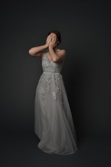 full length portrait of brunette  girl wearing long silver ball gown. standing pose with hands covering face on grey studio background.