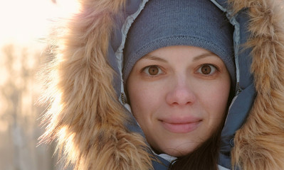 Close-up woman face in fur hood looking in camera and smiling in winter sunny park.