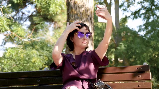 A girl photographs herself by phone, sitting on a bench in the park. Young brunette woman in sunglasses and dress.