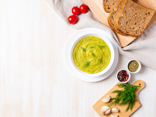 large white bowl with vegetable green cream soup of broccoli, zucchini, green peas on white background, top view, copy space