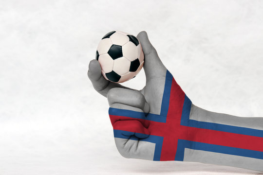 Mini ball of football in Faroe Islands flag painted hand, hold it with two finger on white background. Concept of sport or the game in handle or minor matter.