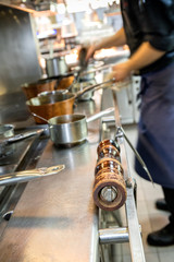 Chef cooking in a commercial kitchen with kitchen utensils in the foreground. A wooden pepper grinder is resting on the stove's side 