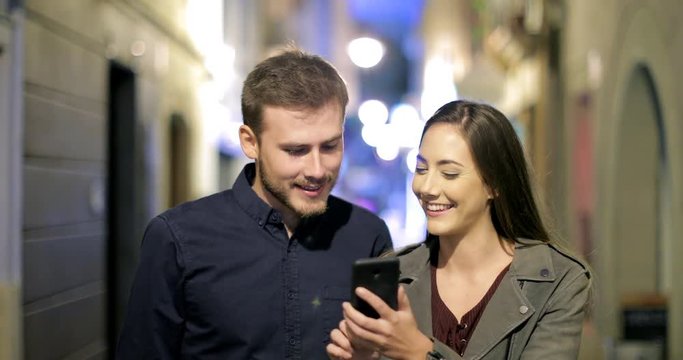 Front view of a happy couple browsing smart phone walking in the night in the street
