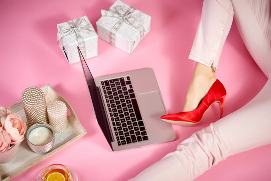 female legs in red high heels, laptop and feminine accessories on a light pink background