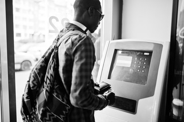 African american man in checkered shirt with backpack holding wallet in hand against atm. Black man...