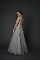 full length portrait of brunette  girl wearing long silver ball gown. standing pose on grey studio background.