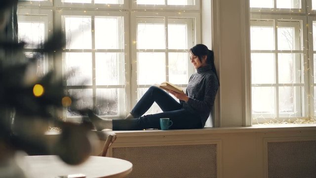 Relaxed girl is reading book on Christmas day sitting on window-sill and looking out of window in December wearing sweater and jeans. Hobby, holiday and youth concept.