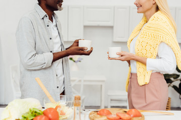 cropped view of mature blonde woman and african american man drinking coffee together at kitchen