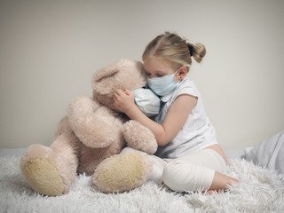 A child in a medical mask on the bed with a big toy bear