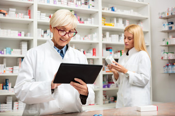 Obraz premium Mature and young female pharmacist working together. Mature woman is using a tablet while young woman reads labels on medication boxes. Medicine, pharmaceutics, health care and people concept