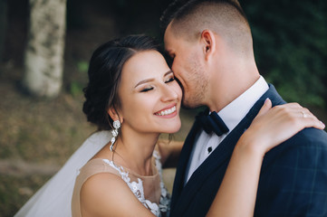 Beautiful newlyweds hugging and smiling in a green garden with trees. Young groom and cute bride close-up. Wedding photography. Stylish portrait.