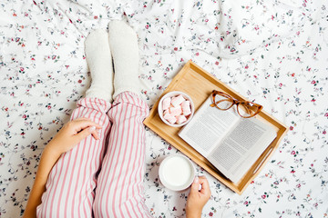 Young woman in pajamas and socks drinking milk at home in bed and reading book, top view