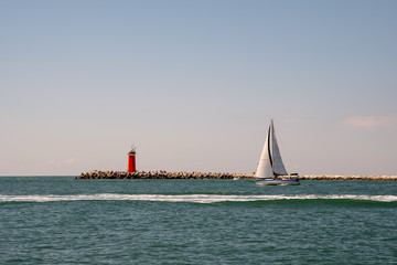 Sailboat in navigation on the Adriatic Sea with a red lighthouse and sunrise sky, Veneto, Italy