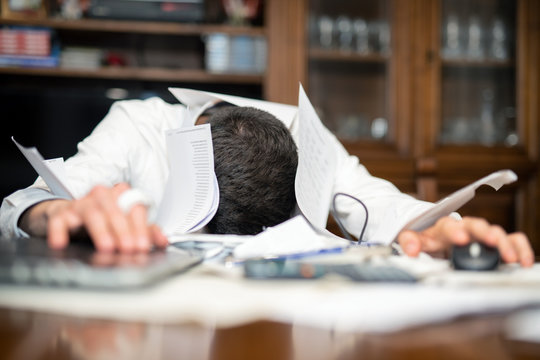 Desperate man submerged from work in his office. Papers and laptop with head on the desktop