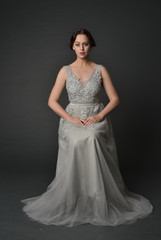 Fototapeta premium full length portrait of brunette girl wearing long silver ball gown. seated pose on grey studio background.