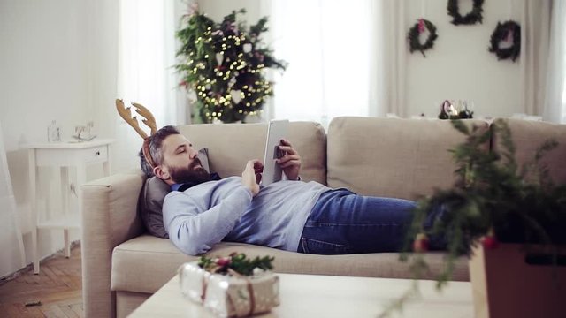 A man with reindeer headband lying on a sofa at Christmas time, using tablet.