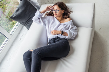young pretty sexy charming girl in a white man's shirt lying on a white sofa near the window in the...
